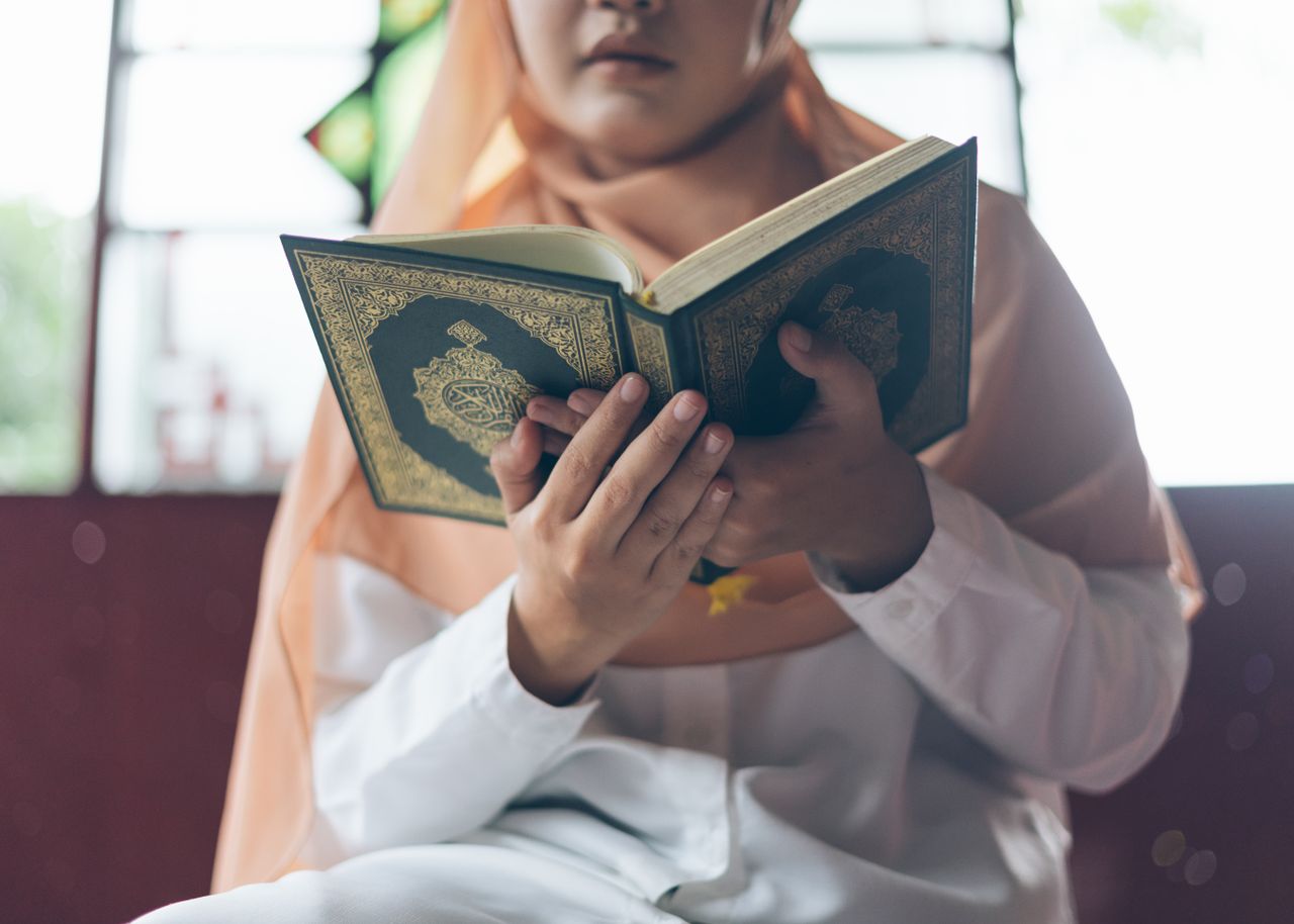 A Muslim woman in a hijab reads the Quran indoors, capturing a spiritual moment.